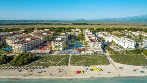 An aerial view of a coastal resort with multiple buildings, a sandy beach, and clear blue waters. - Grupotel Alcudia Pins (Muro)