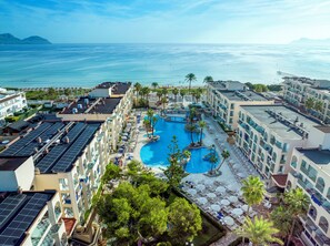 A resort with a large pool surrounded by palm trees and sun loungers, overlooking the ocean.