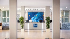 A modern reception area with a blue mural, two potted plants, and a sign reading "BLUE Acadia Pines." - Grupotel Alcudia Pins (Muro)