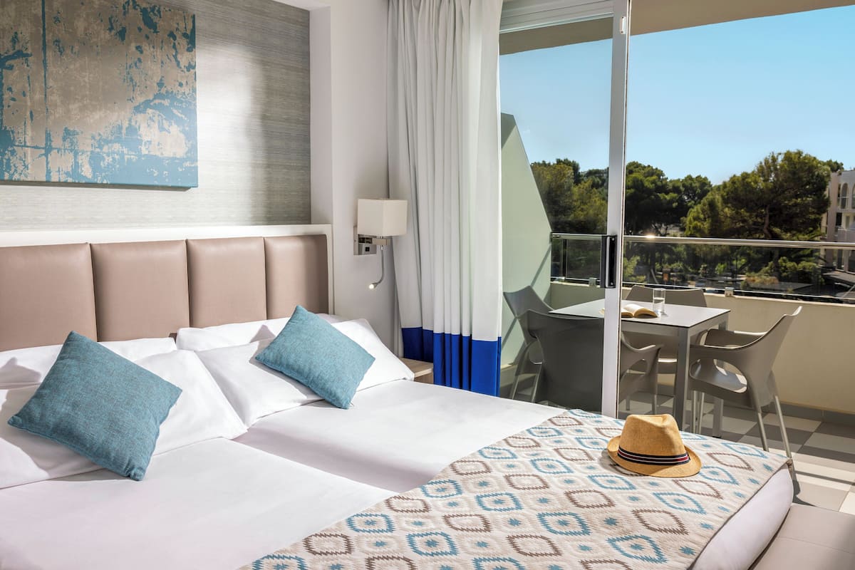 A hotel room with a large bed, a patterned throw blanket, a hat on the bed, a desk with a chair, and a balcony with a view of trees and buildings.