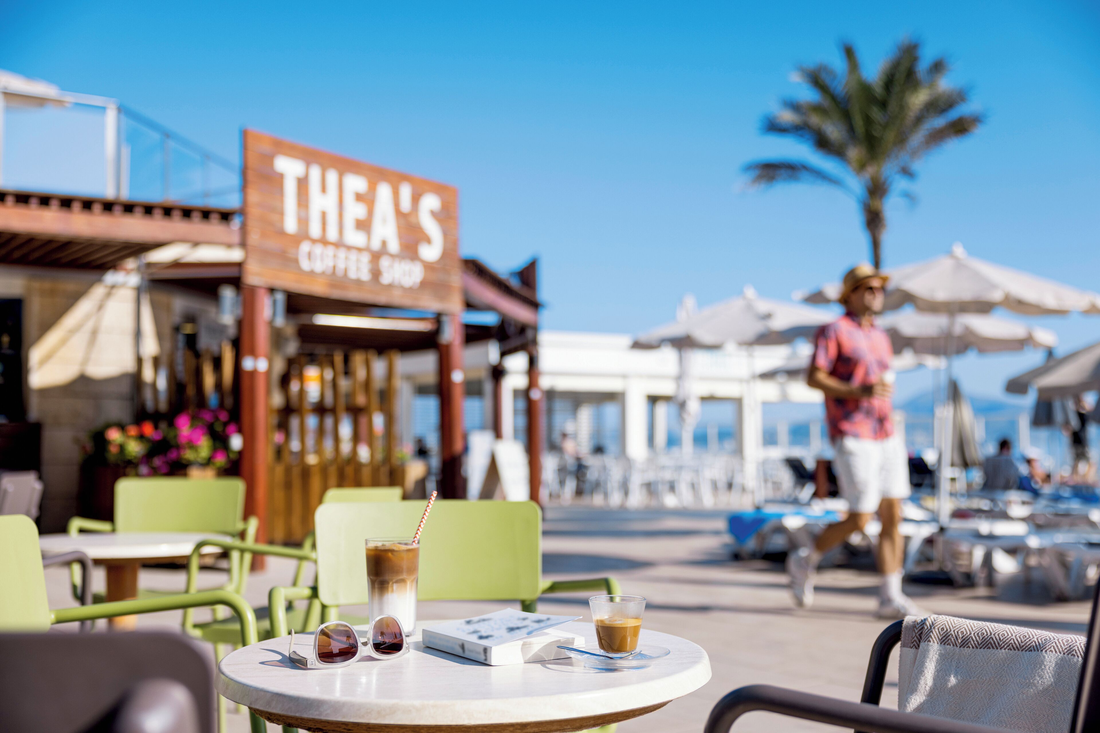 En café med udendørs siddepladser, et skilt med teksten "THEA'S COFFEE SHOP" og et palmetræ i baggrunden.