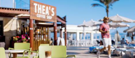 A cafe with outdoor seating, a sign reading "THEA'S COFFEE SHOP", and a palm tree in the background.