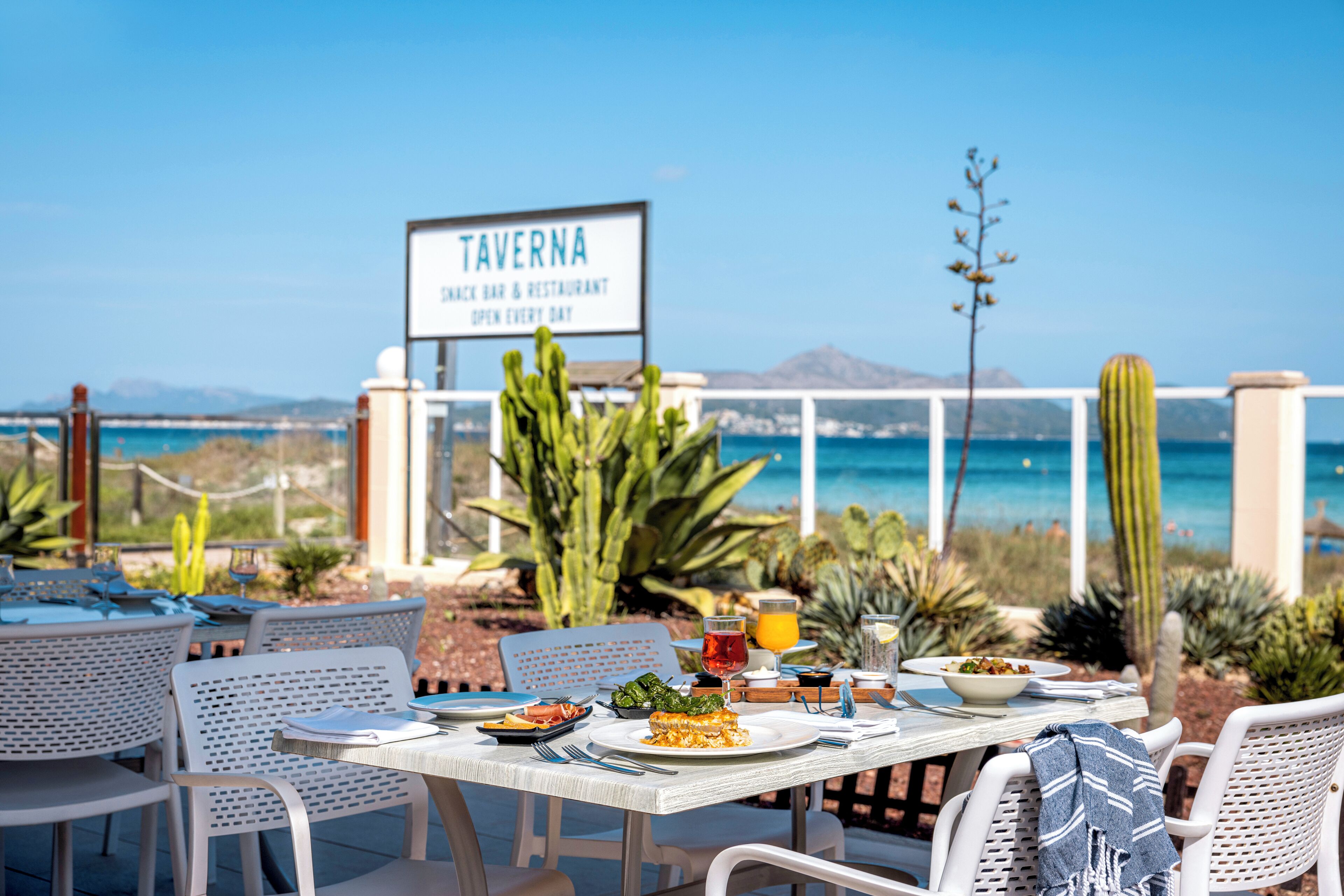 Ein Essbereich im Freien mit Tischen und Stühlen, ein Schild für das Restaurant 'TAVERNIA' und im Hintergrund Meer und Berge.