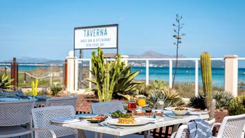 Ein Essbereich im Freien mit Tischen und Stühlen, ein Schild für das Restaurant 'TAVERNIA' und im Hintergrund Meer und Berge.