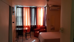 Une petite pièce bien éclairée, avec un lit, un bureau et une chaise, ainsi qu’un fauteuil rouge.