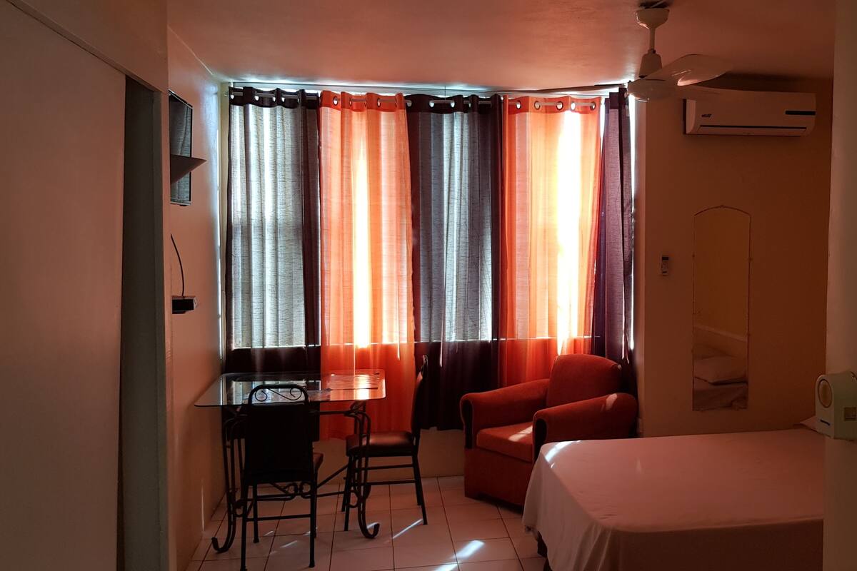 A small, well-lit room with a bed, a desk with a chair, and a red armchair.