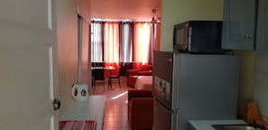 Compact kitchen with refrigerator, microwave, sink, and kettle. Dining area with table and chairs. Sofa near the window.