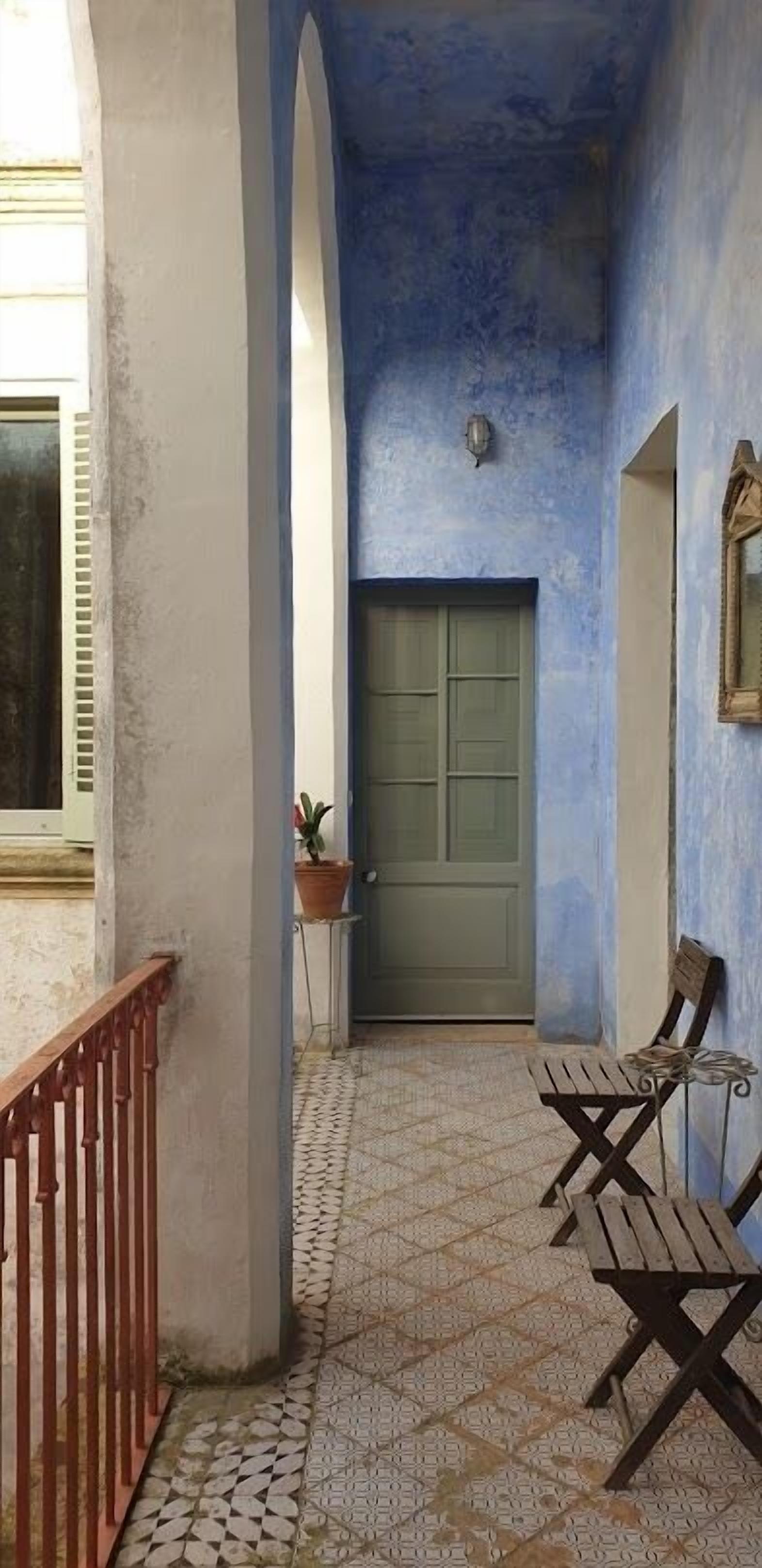 Um corredor com piso de azulejos, uma porta verde e duas cadeiras de madeira.