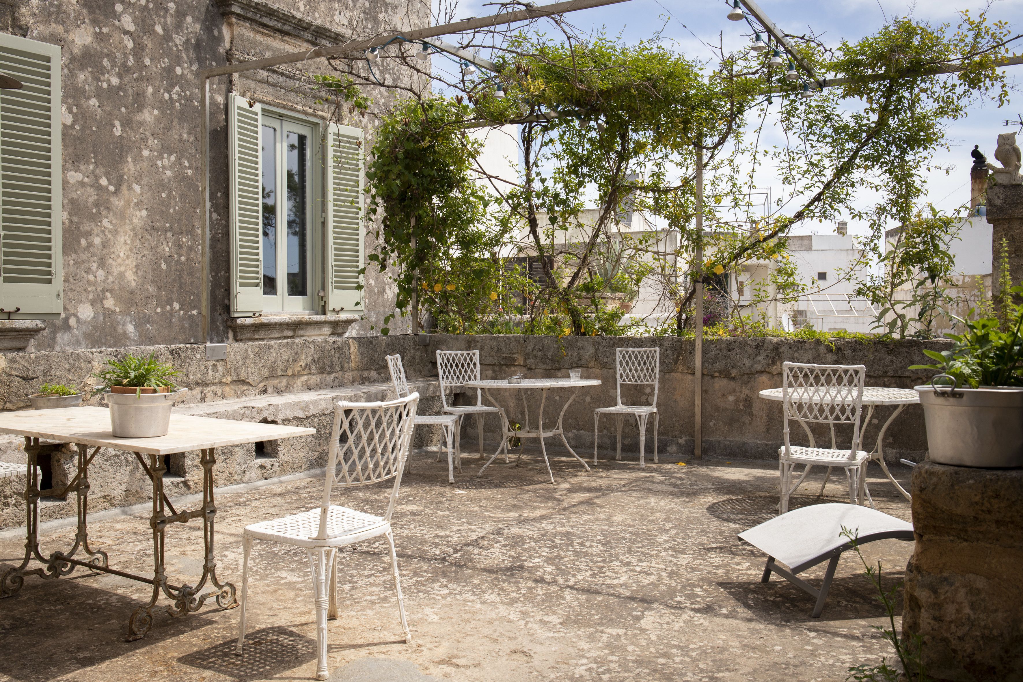 An outdoor patio with white metal furniture, including tables and chairs, surrounded by stone walls and greenery.