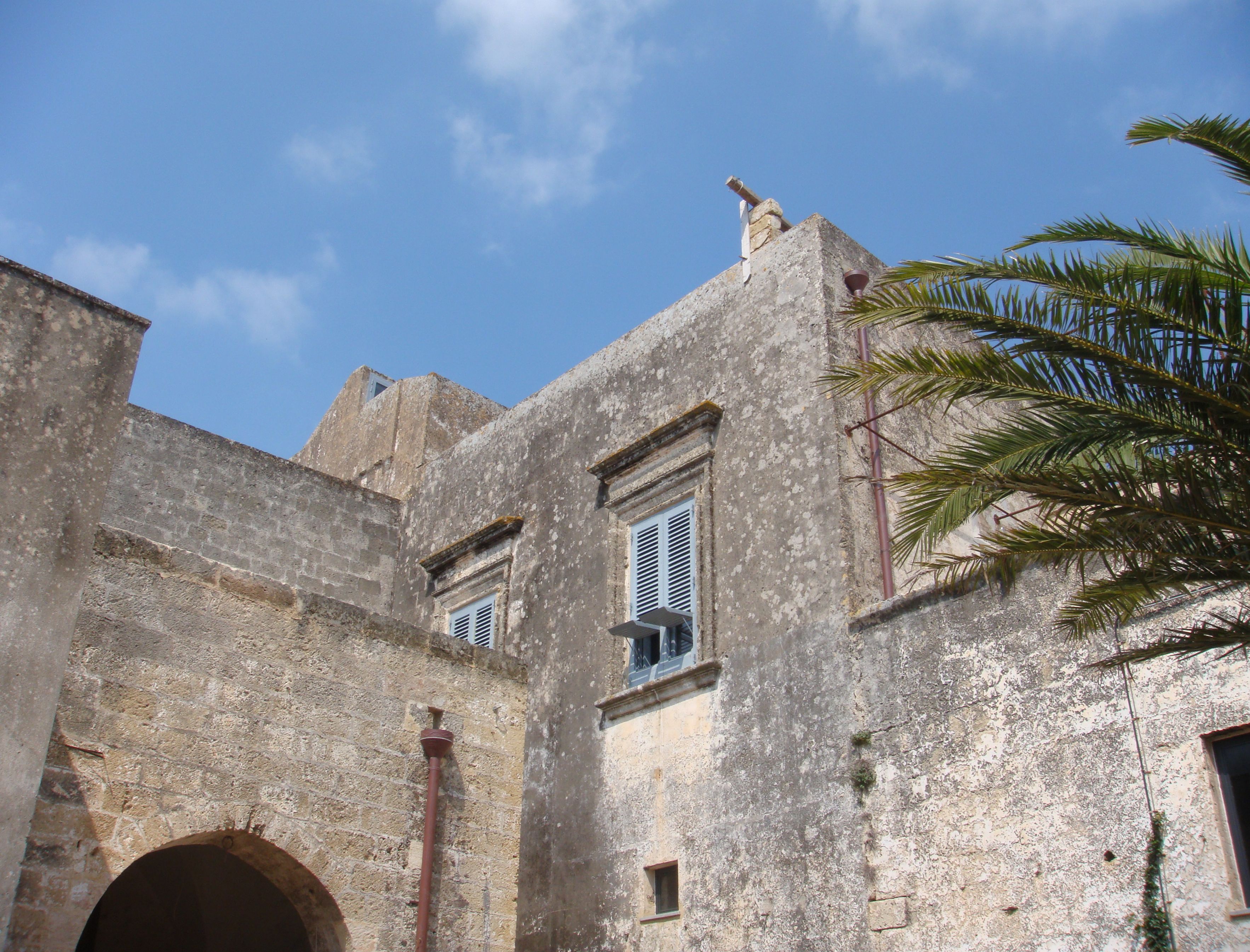 A historic building with stone walls, a small window with shutters, and a palm tree in the foreground.