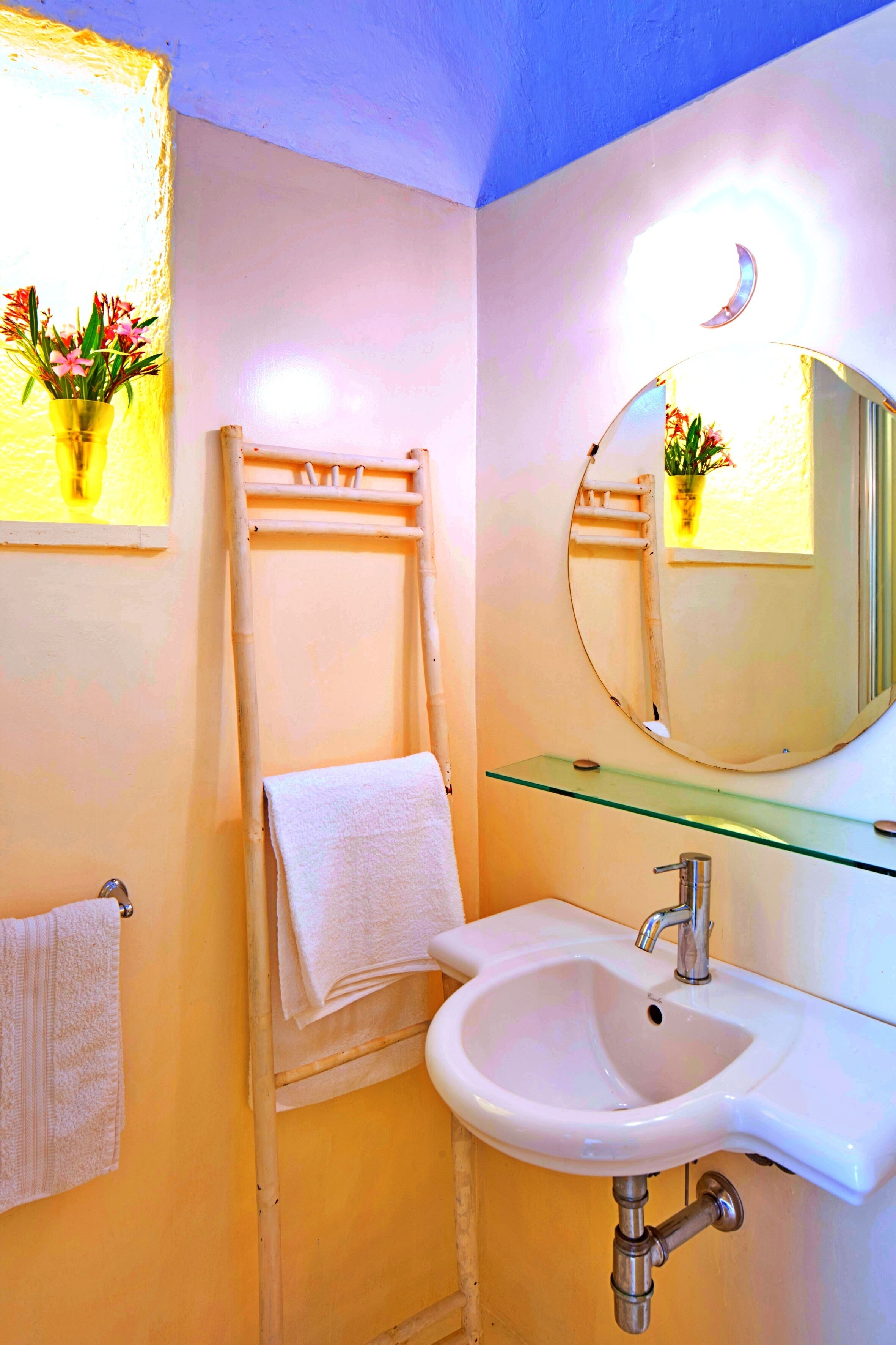 A bathroom with a white sink, a glass shelf, a towel rack, and a mirror with a crescent moon light fixture.