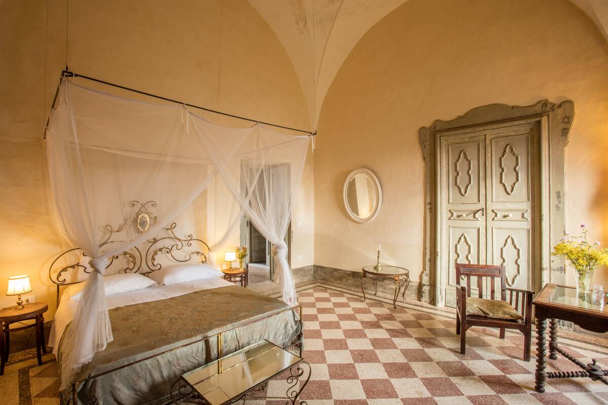 Ein Zimmer mit einem Himmelsbett, einem Holzstuhl, einem Tisch mit einer Vase und einer Tür mit dekorativen Feldern.