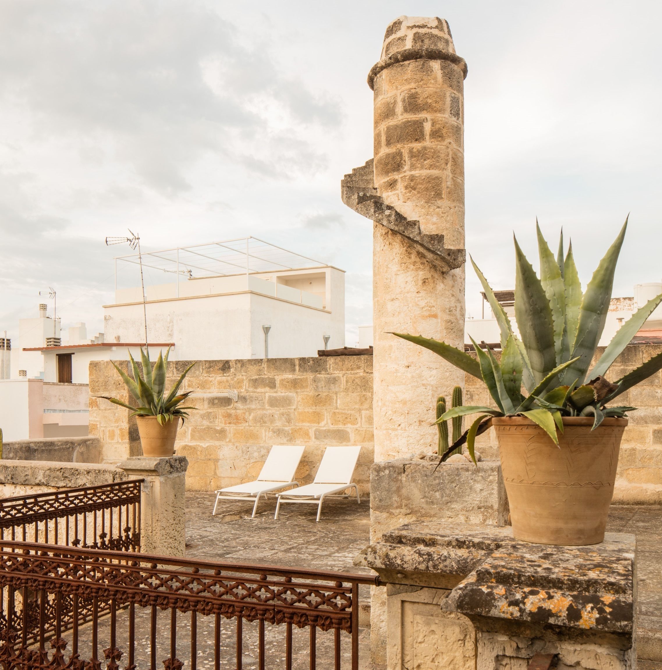 A rooftop terrace with stone walls, two potted aloe plants, and two white lounge chairs.