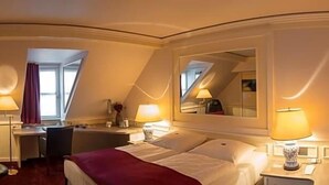 A hotel room with a bed, desk, chair, lamps, and a mirror.