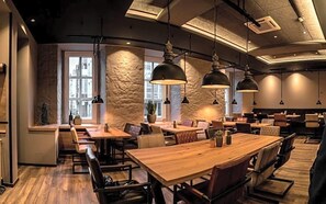 Interior de restaurante con mesas y sillas de madera, iluminación colgante y una pared decorativa de piedra.