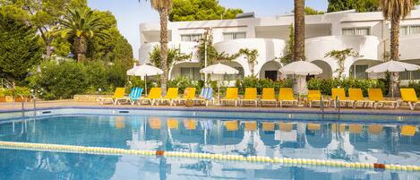A clear swimming pool with yellow lounge chairs and umbrellas, surrounded by palm trees and a building with multiple balconies.