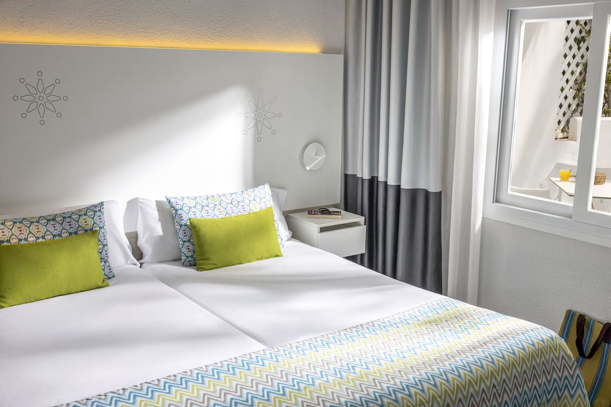 A neatly made bed with a patterned bedspread, a bedside table with a lamp, and a window with curtains.