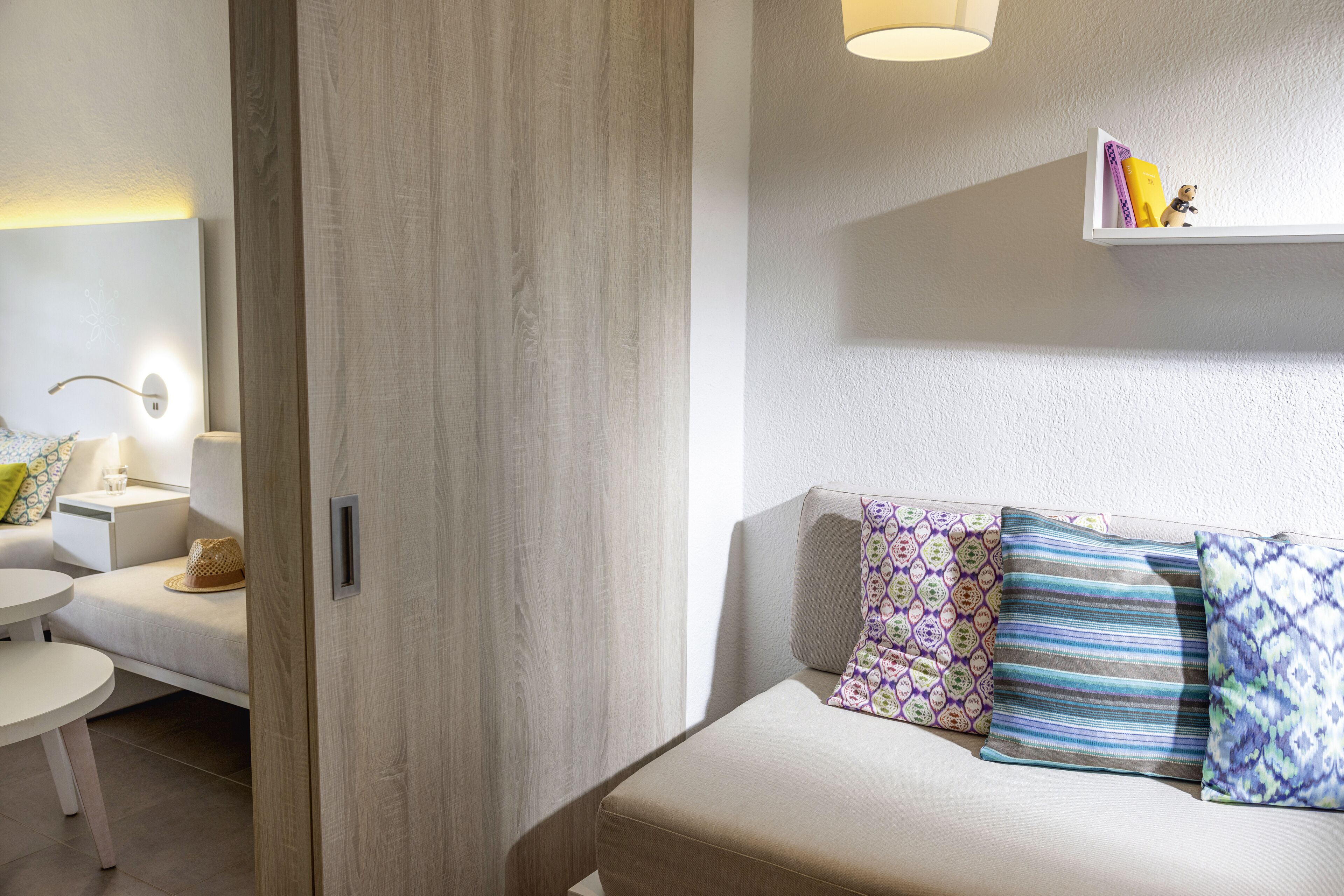 A modern room with a wooden door, a white sofa with patterned cushions, a small table, and a shelf with books and a figurine.