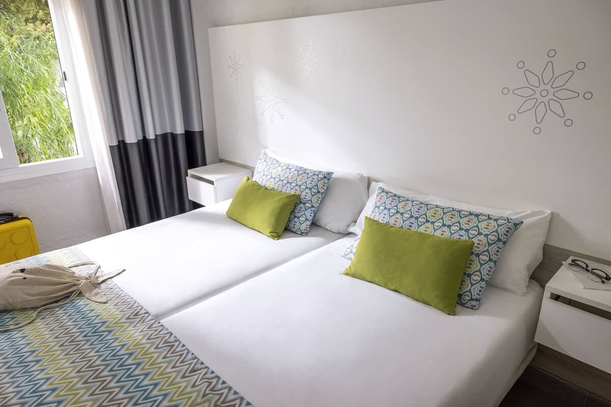 A hotel room with two beds, a window with curtains, a bedside table with a lamp, and a wall with decorative patterns.