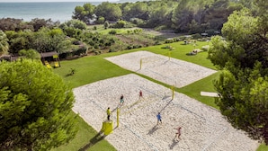 A beach volleyball court with a net, surrounded by green grass and trees, with a view of the sea and mountains in the background.