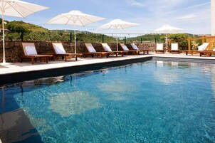 Zona de piscina con sillones y sombrillas, una pared de piedra y vistas a las colinas.