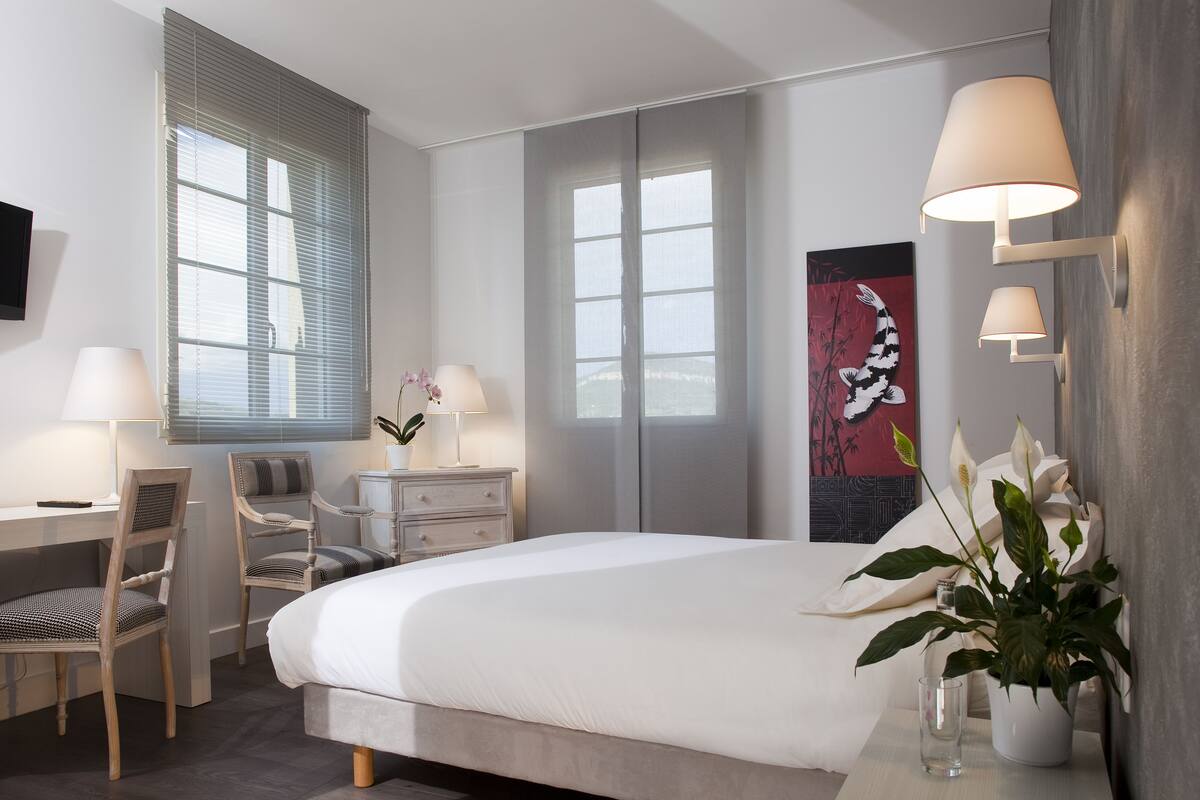 A modern hotel room with a large bed, a desk with a chair, a bedside table with a lamp, and a large window with blinds.