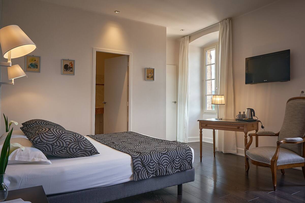 A hotel room with a bed, a desk with a chair, a TV, and a window with curtains.