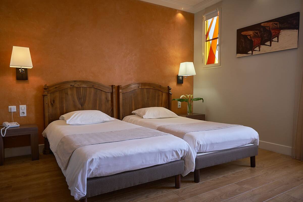A hotel room with two beds, a wooden headboard, bedside lamps, a small table, and a painting on the wall.