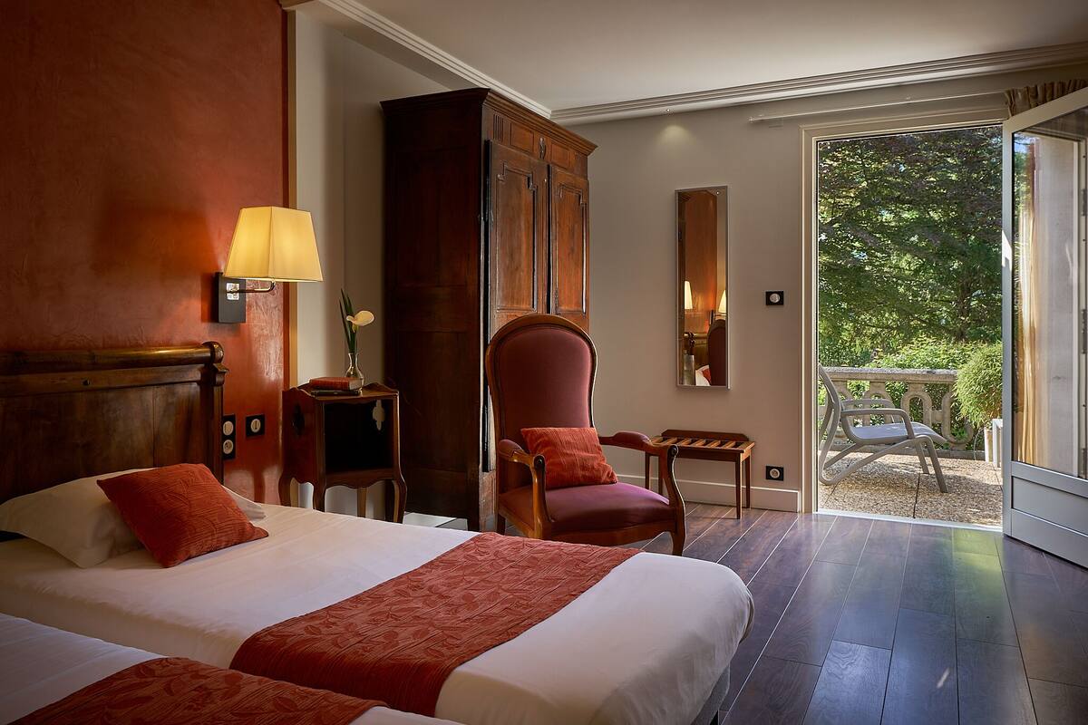 A hotel room with two beds, a wooden wardrobe, a red armchair, a small table, and a view of a garden through a glass door.