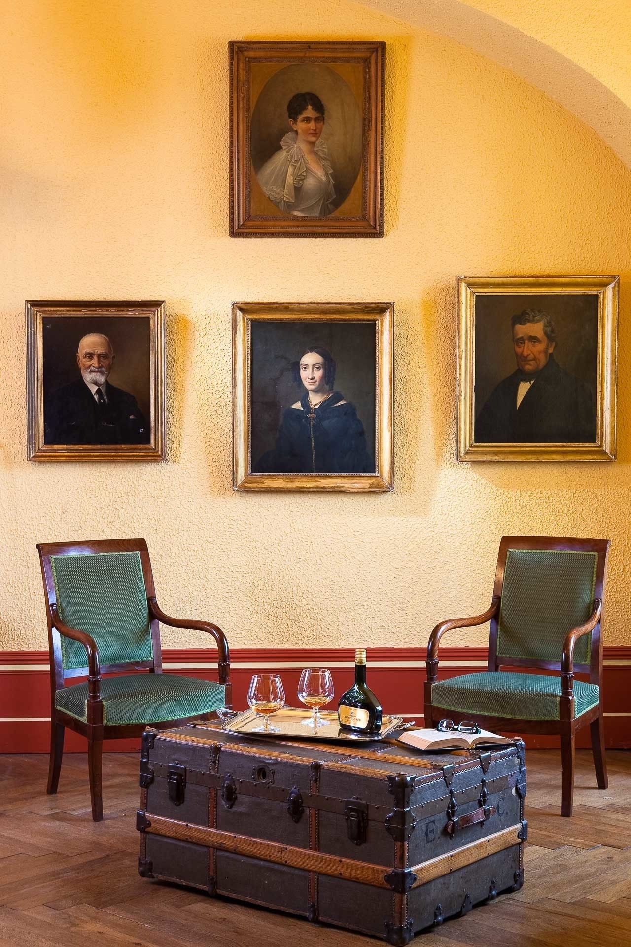 Een kamer met drie ingelijste portretten aan de muur, twee groene bekleden stoelen, een houten tafel met een dienblad met glazen en een fles, en een vintage koffer.