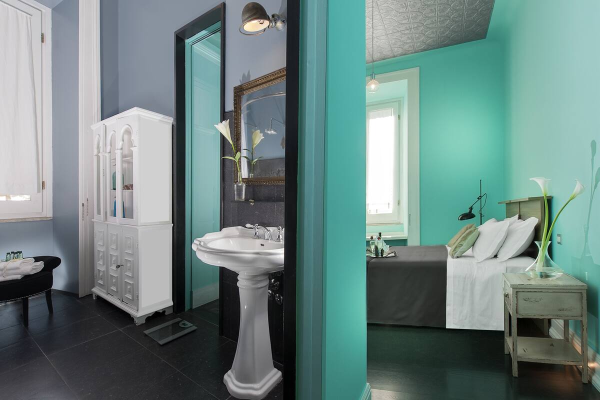 A bathroom with a pedestal sink, a white cabinet, and a bed with a teal wall.