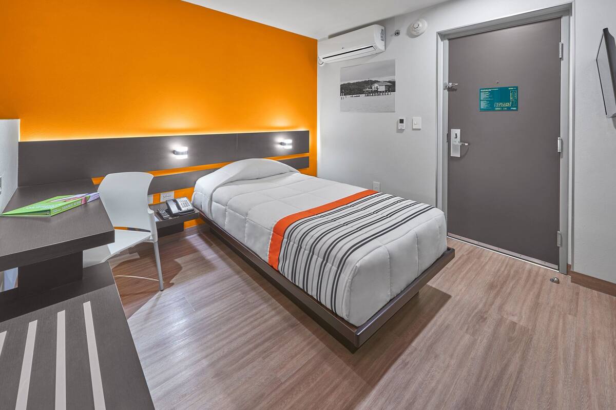 A hotel room with a bed, a desk, and a chair. There is a door with a sign on it, and an air conditioning unit on the wall.