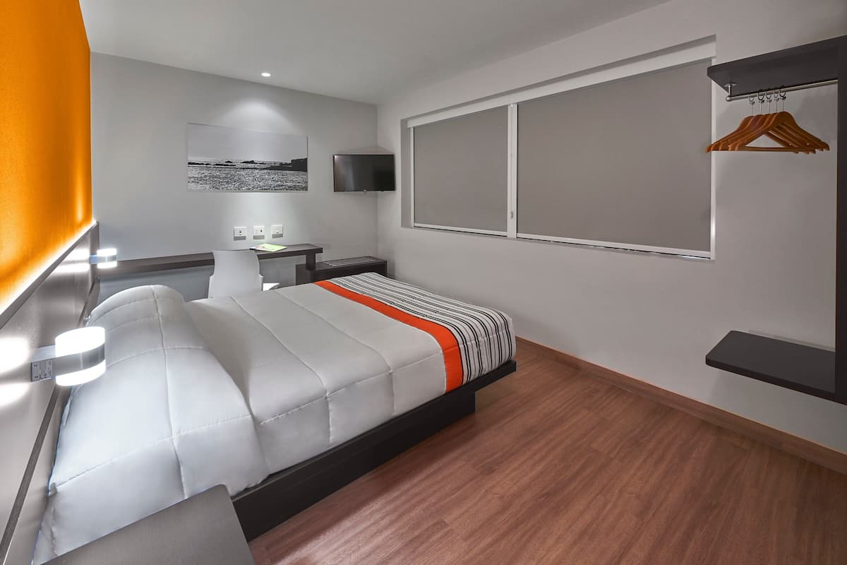A modern hotel room with a large bed, bedside tables, a flat-screen TV, a window with blinds, and a wall-mounted TV.