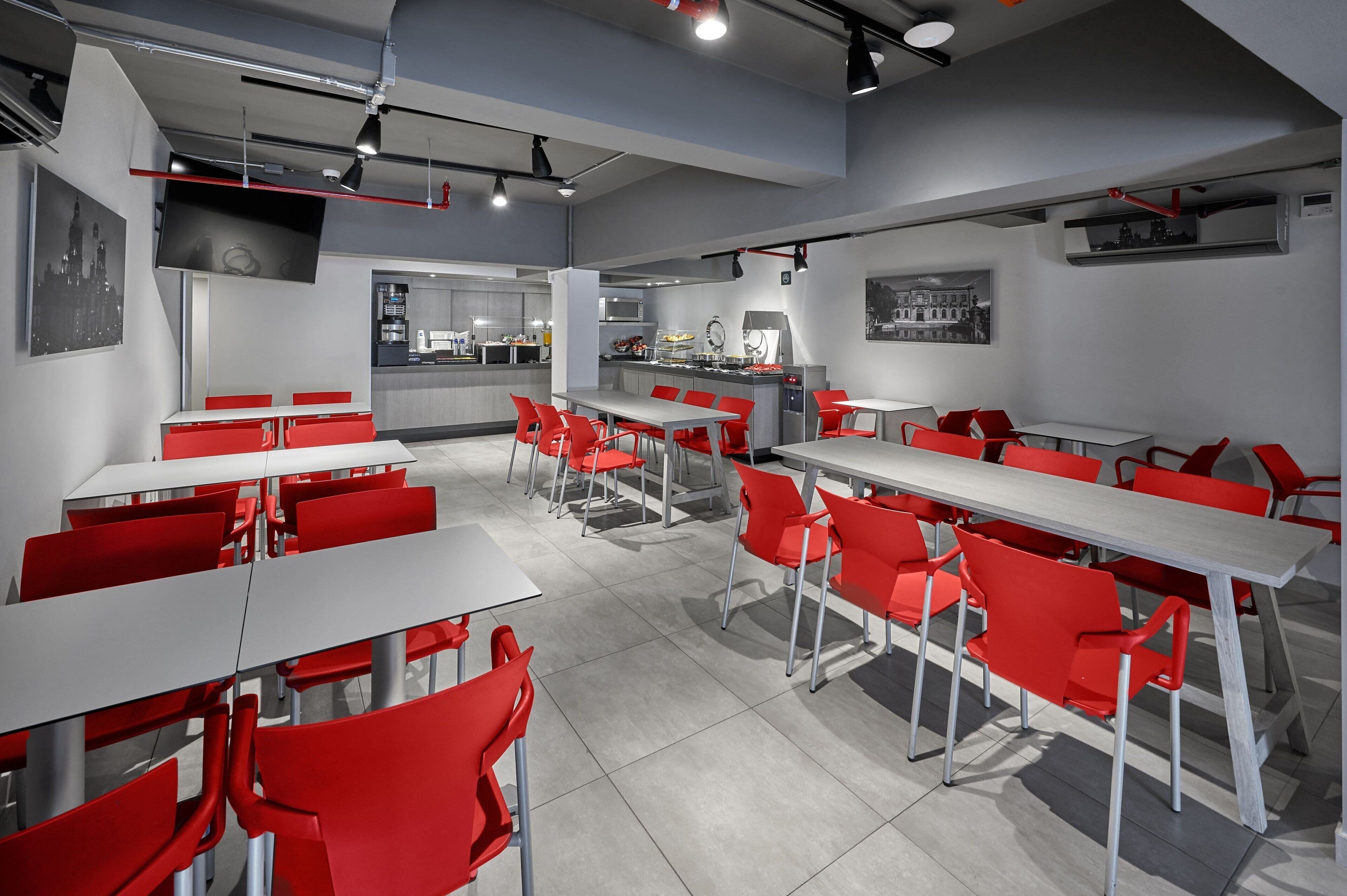 A modern restaurant with red chairs and white tables, a kitchen area, and cityscape artwork on the walls.