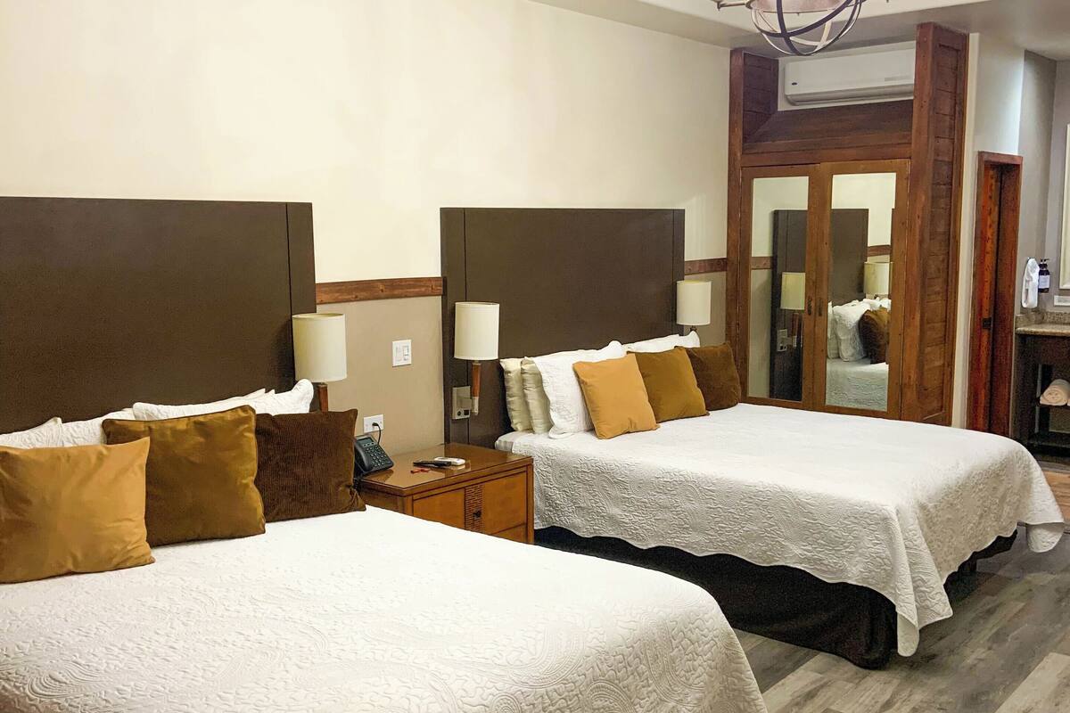Une chambre d’hôtel avec deux lits, chacun recouvert d’une couvre-lit blanche et agrémenté d’oreillers marron. On y trouve une tête de lit en bois, une table de chevet avec une lampe et un lustre.