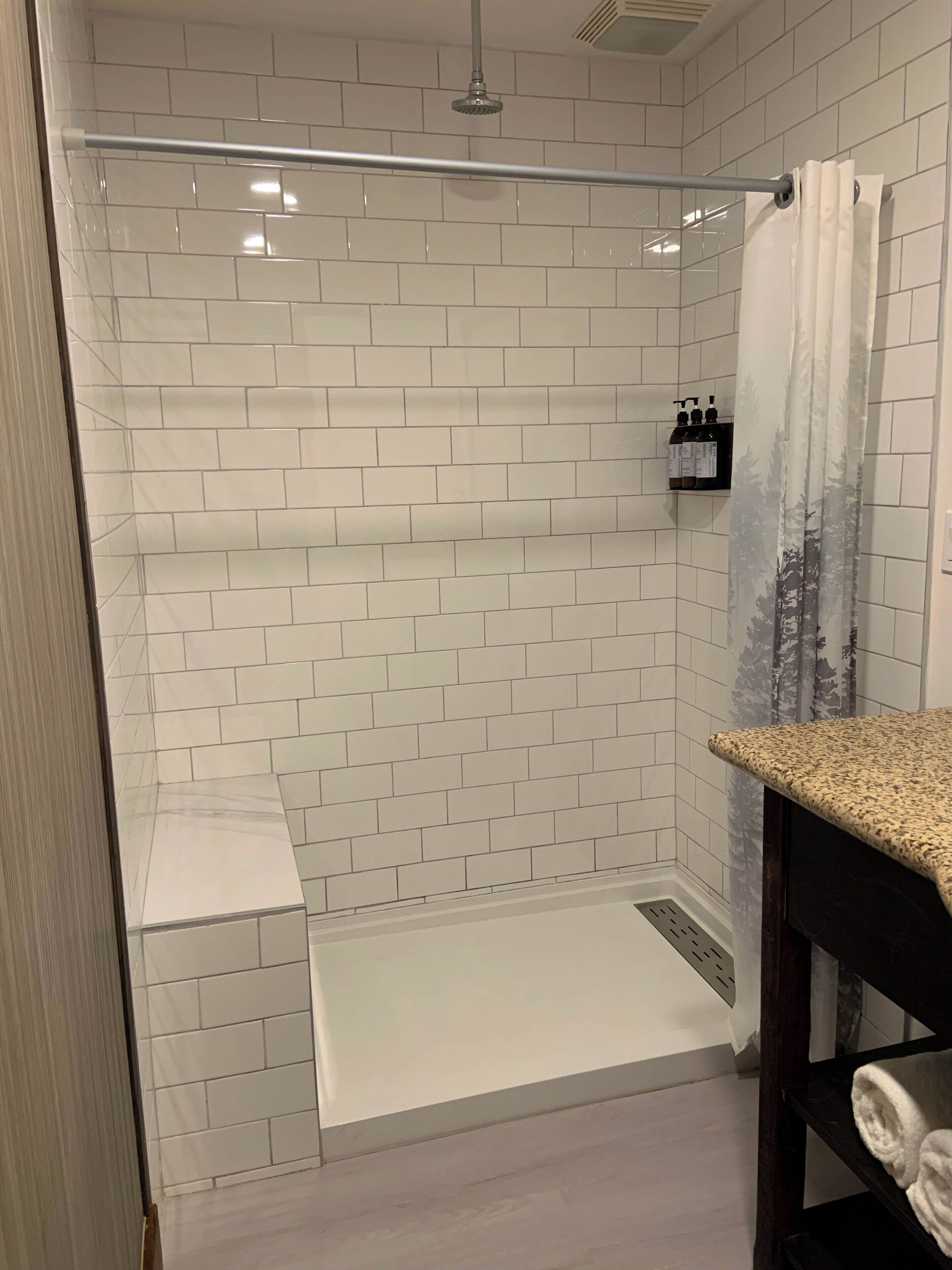 Een moderne badkamer met witte metrotegels, een glazen douchecabine, een houten wastafelmeubel met een handdoek en een klein plankje met toiletartikelen.