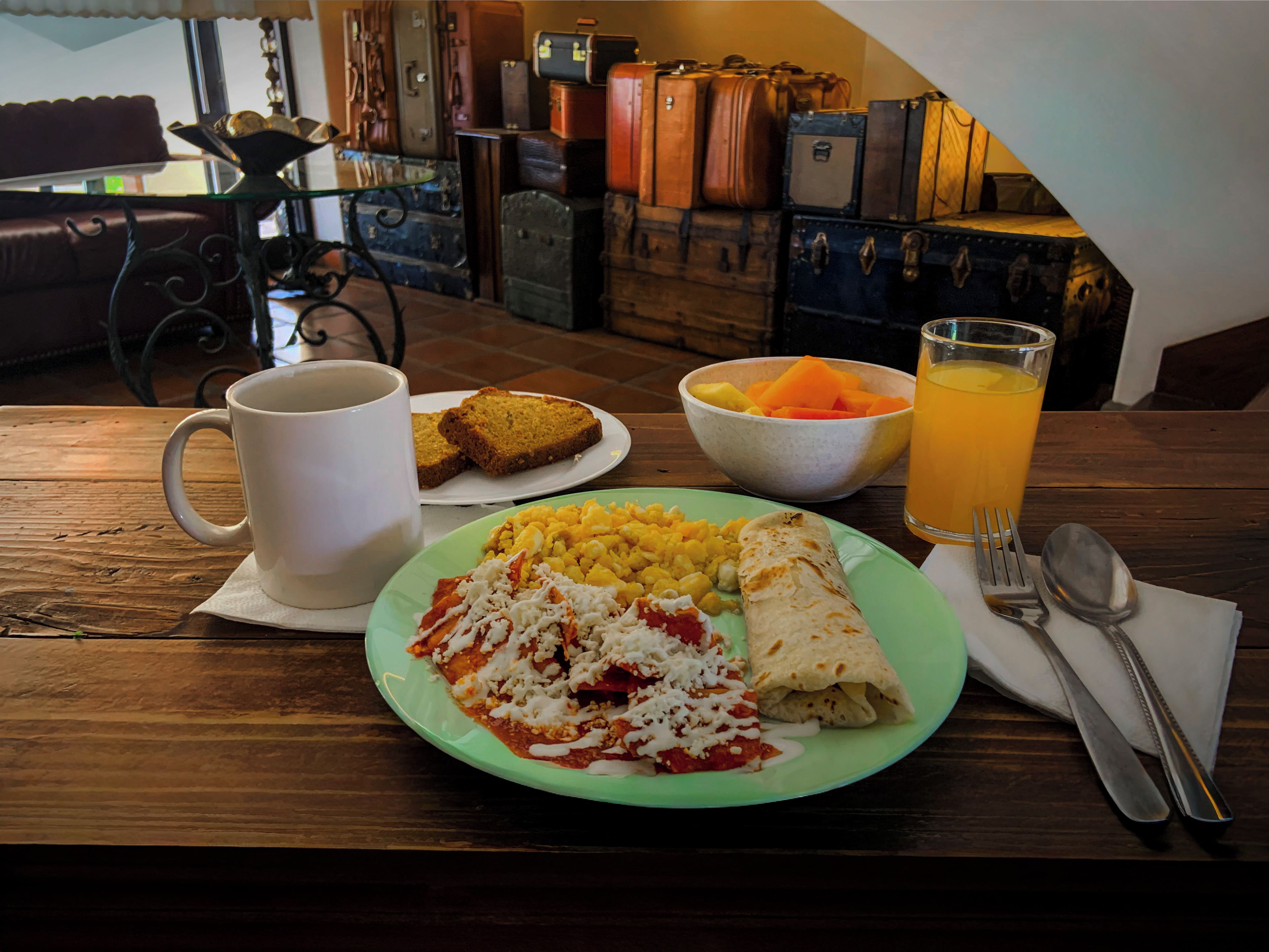 A breakfast plate with eggs, toast, and a burrito, a mug, a bowl of fruit, and a glass of orange juice on a wooden table.