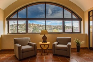 A room with two patterned armchairs, a small round table with a lamp, and a large arched window offering a view of a hilly landscape.