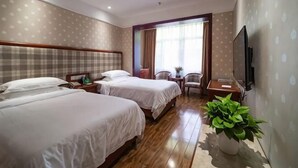 Una habitación de hotel con dos camas, televisión, escritorio, silla, ventana con cortinas y una planta en maceta.