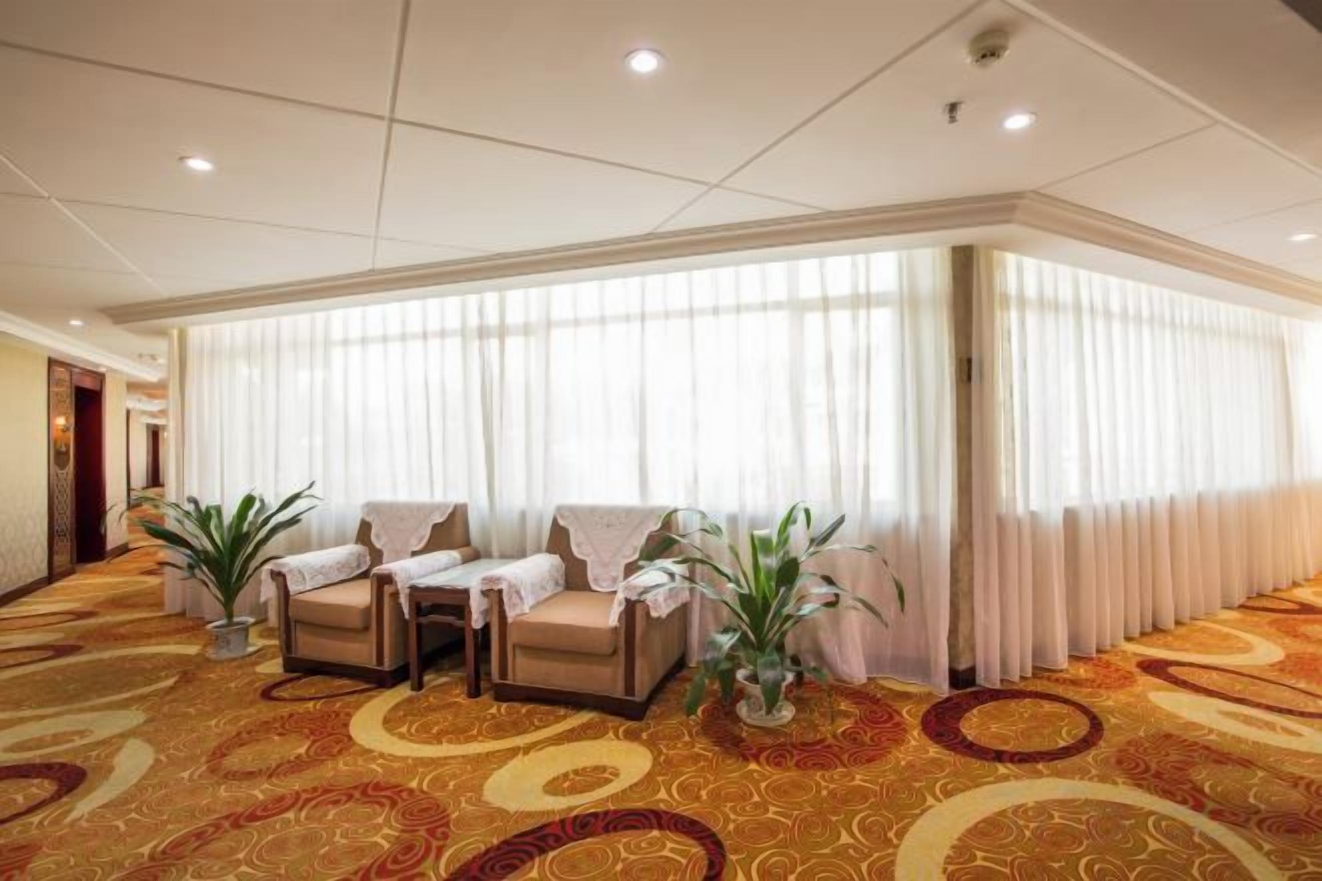 A hotel lobby with a carpeted floor, a seating area with chairs and a table, and a large window with curtains.