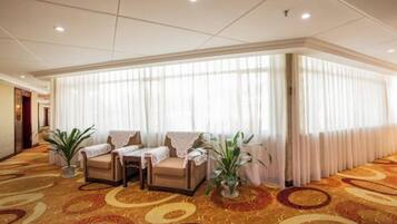 A hotel lobby with a carpeted floor, a seating area with chairs and a table, and a large window with curtains.