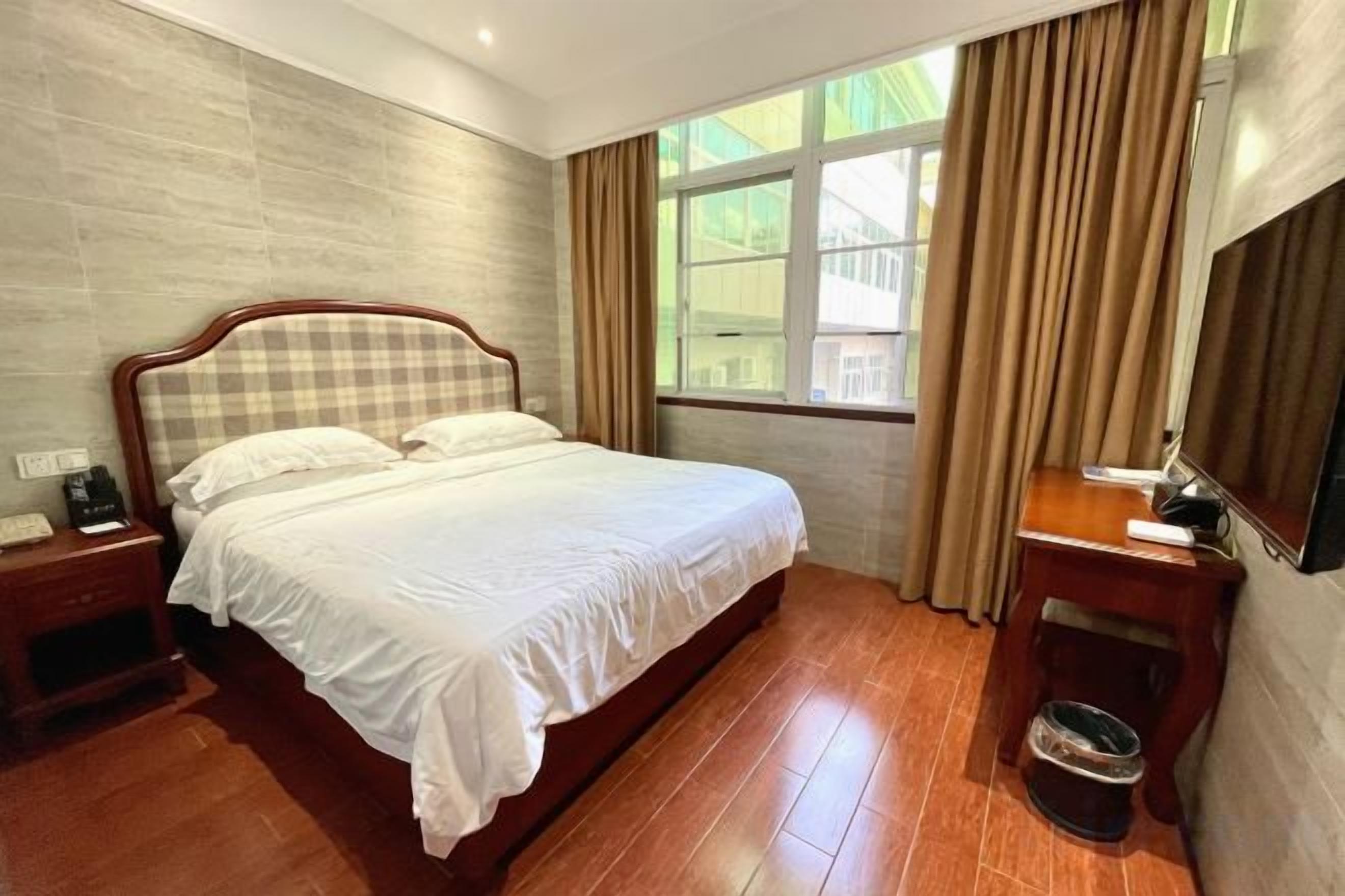 A hotel room with a large bed, a bedside table with a lamp, a small table with a chair, a window with curtains, and a wall-mounted TV.
