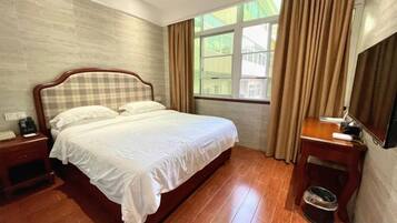 A hotel room with a large bed, a bedside table with a lamp, a small table with a chair, a window with curtains, and a wall-mounted TV.