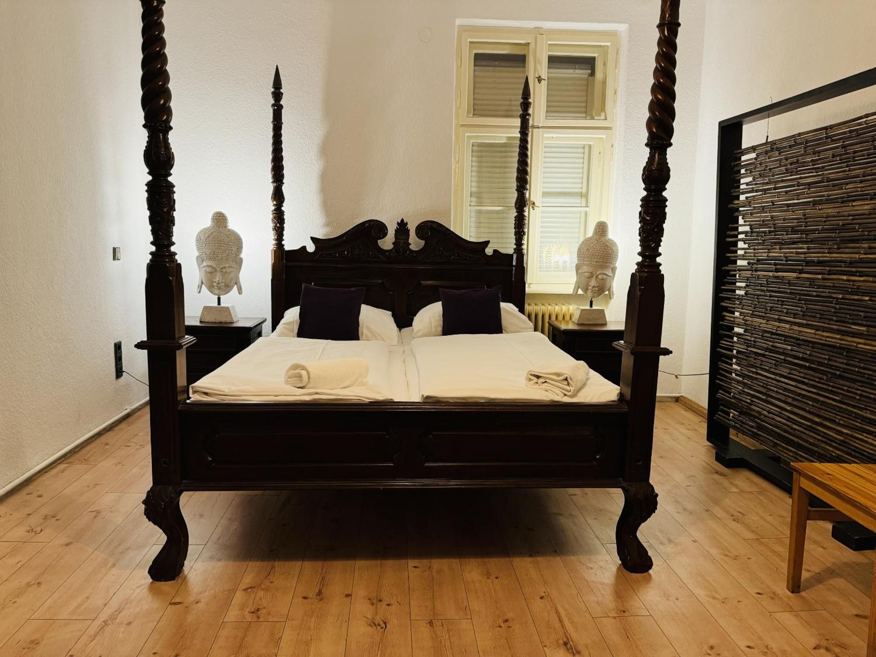 Una cama con dosel de acabado oscuro, cabecera blanca y una ventana con contraventanas al fondo.