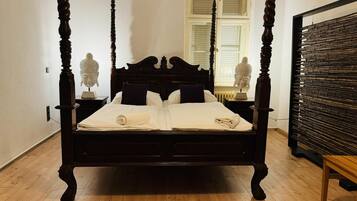 Una cama con dosel de acabado oscuro, cabecera blanca y una ventana con contraventanas al fondo.