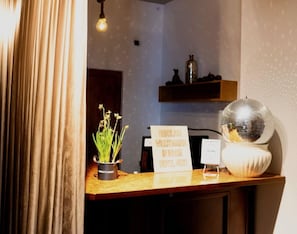 Un mostrador de recepción de hotel con un cartel de bienvenida, una planta en maceta, una bola de discoteca y una estantería con adornos.