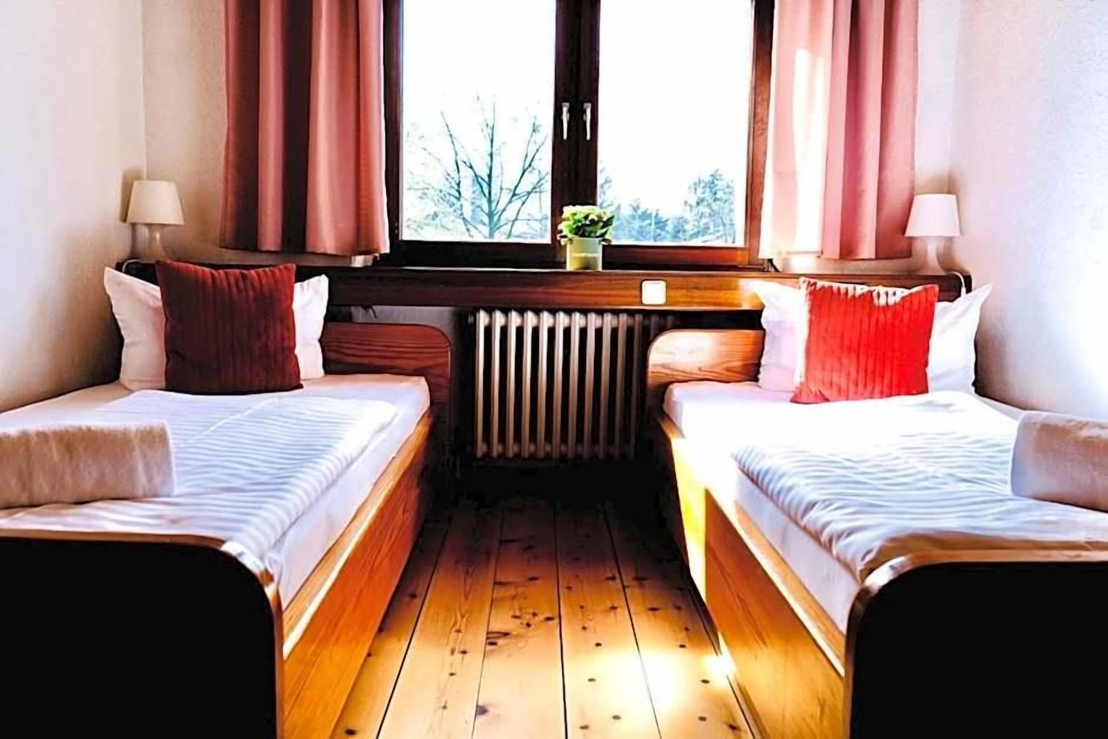 Ein Zimmer mit zwei Einzelbetten, einem Heizkörper, einem Fenster mit Vorhängen und einer kleinen Pflanze auf der Fensterbank.