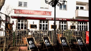 Ein Sitzbereich im Freien mit Tischen und Stühlen, ein Schild für 'Fish & Beef Club' und ein Gebäude mit zahlreichen Fenstern im Hintergrund.