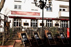 An outdoor seating area with tables and chairs, a sign for 'Fish & Beef Club', and a building with multiple windows in the background. - Hotel Nest (Berlin)
