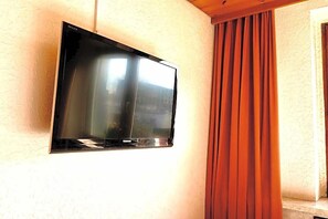 A flat-screen TV mounted on a textured wall, a window with curtains, and a wooden ceiling beam. - Hotel Nest (Berlin)